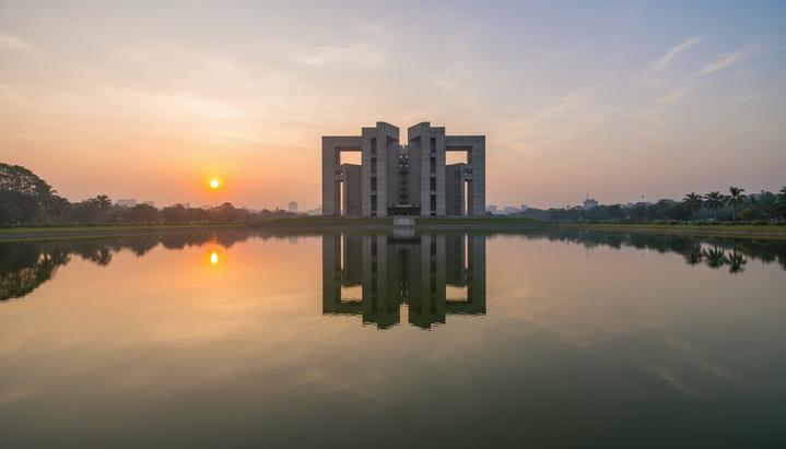 Здание Национального парламента Бангладеш в Дакке, отражающееся в озере на рассвете. Архитектурный комплекс на берегу водоема.