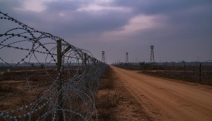 Забор из колючей проволоки на границе Индии и Бангладеш на фоне пасмурного закатного неба и отдаленных сторожевых вышек.