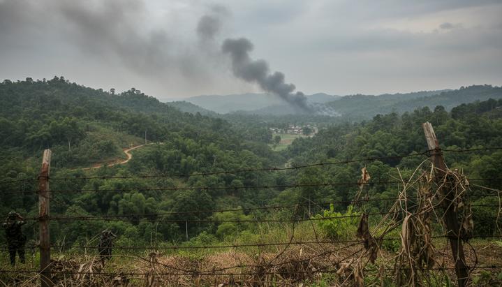 Бангладеш в тисках соседей: Индия и Мьянма испытывают Дакку на прочность Забор из колючей проволоки на границе Бангладеш и Мьянмы, за которым видны зеленые холмы и дым от далекого пожара.