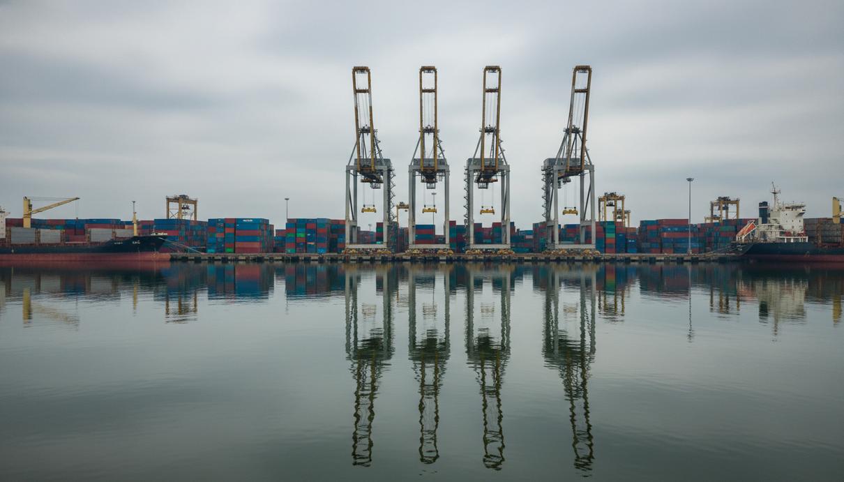 Ряды разноцветных морских контейнеров в торговом порту на фоне массивных кранов и пасмурного неба